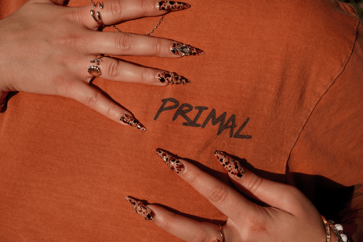 Close-up of a person's hands with long leopard-print manicured nails resting on an orange t-shirt that has the word 'PRIMAL' printed on it.