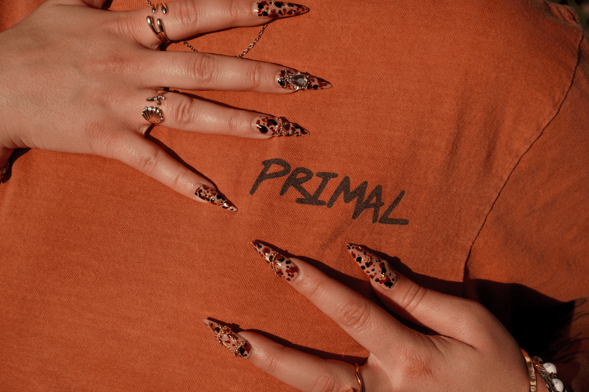 Close-up of a person's hands with long leopard-print manicured nails resting on an orange t-shirt that has the word 'PRIMAL' printed on it.